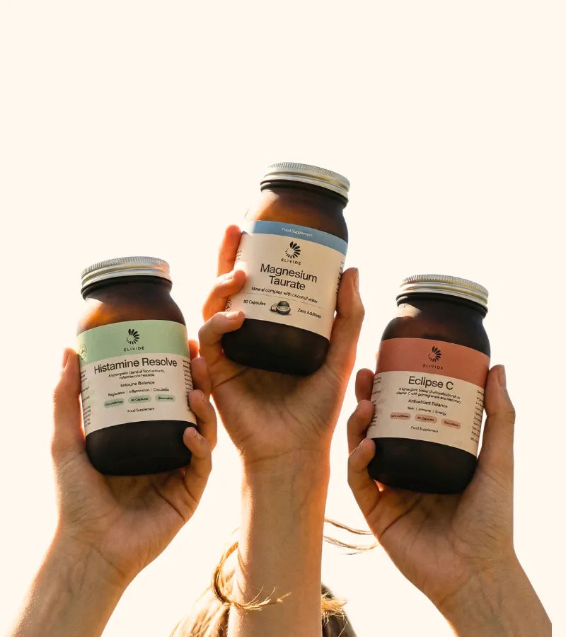 Three hands holding ELIVIDE supplement bottles against a light sky background